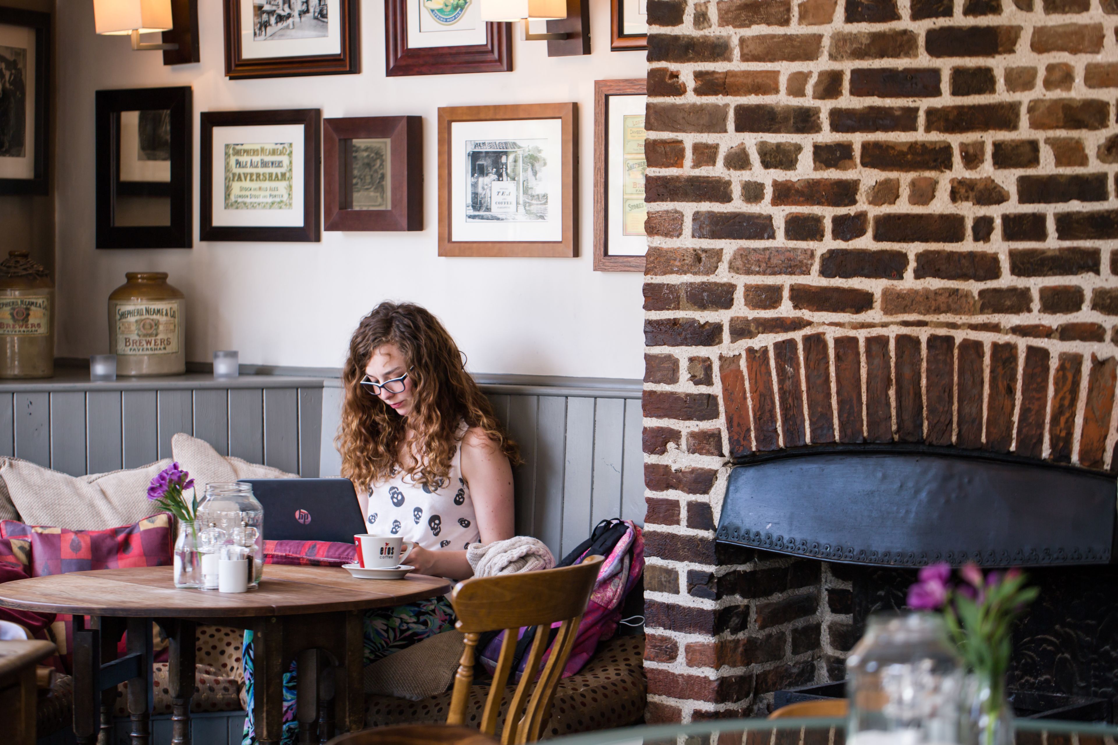 A cozy cafe with a brick fireplace, framed pictures on the wall, and a table with a laptop and a cup.