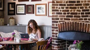 A cozy cafe with a brick fireplace, framed pictures on the wall, and a table with a laptop and a cup.
