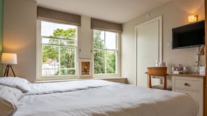 A hotel room with a large bed, a desk, a chair, a TV, and a window with a view of trees and buildings. - The Millers Arms (Canterbury)