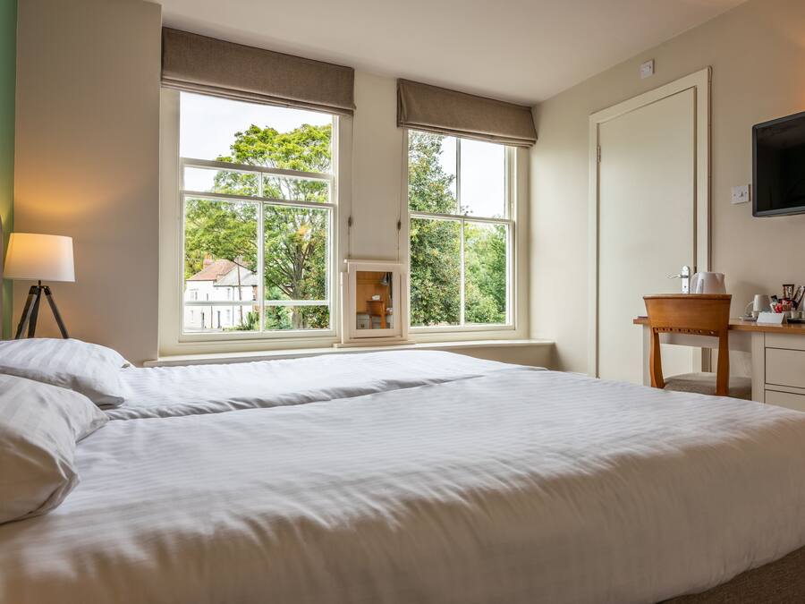 A hotel room with a large bed, a desk, a chair, a TV, and a window with a view of trees and buildings.