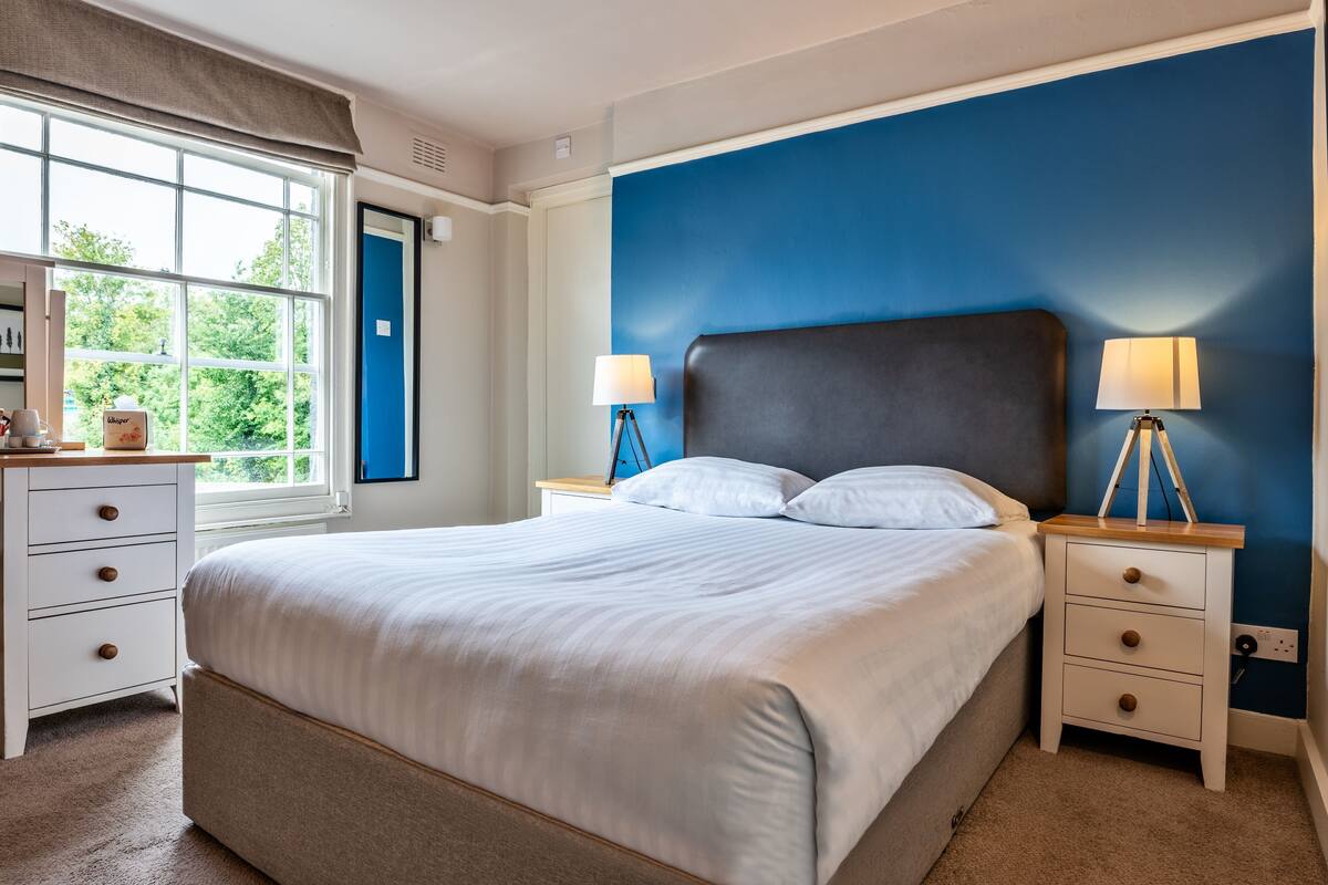 A bedroom with a large bed, two bedside tables with lamps, a dresser, and a window with a view of greenery.