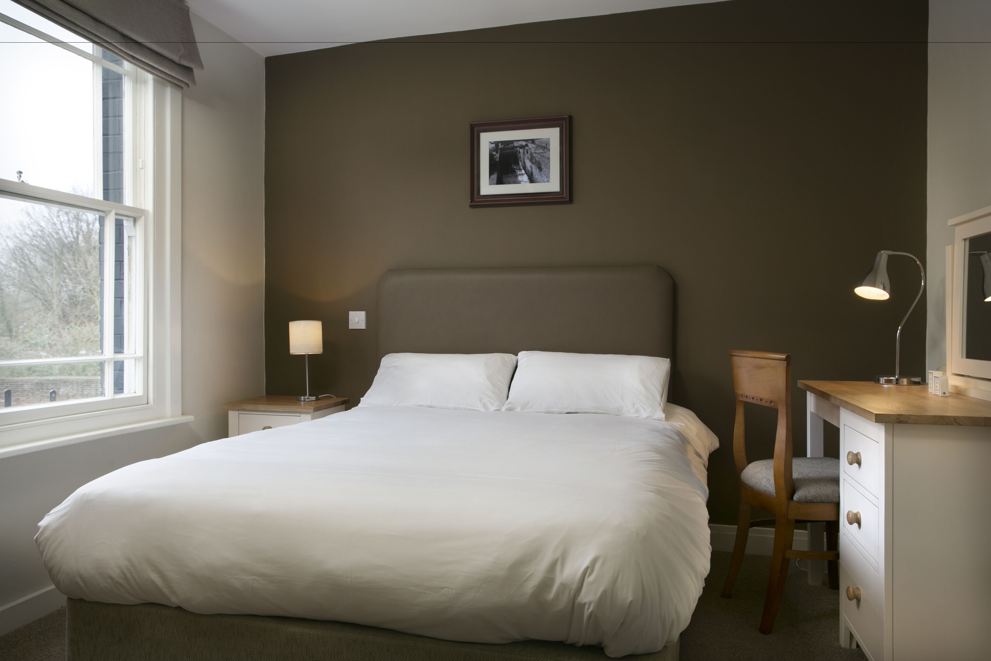 A neatly made bed in a room with a large window, a bedside table with a lamp, a chair, and a dresser with drawers.