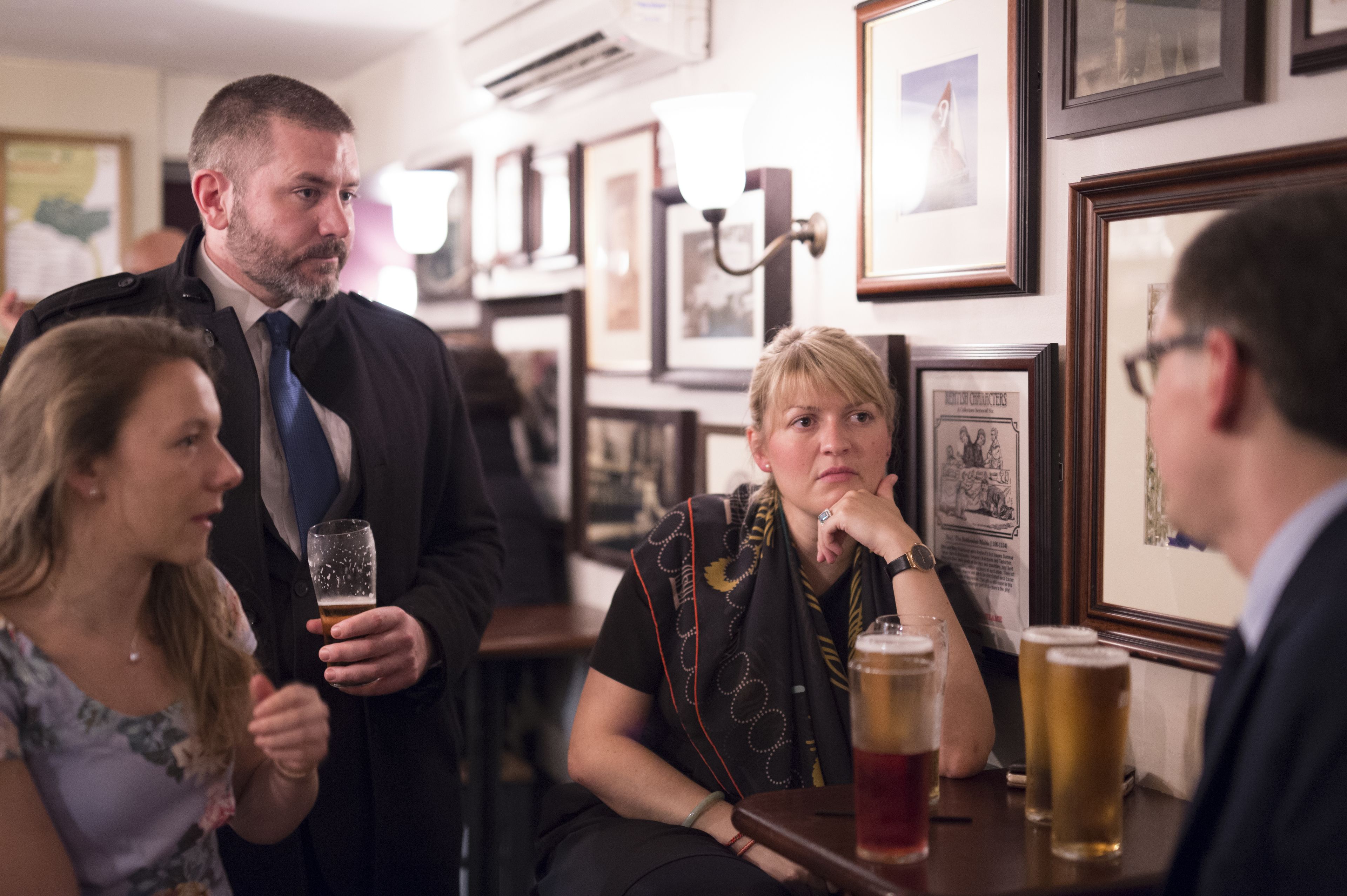 A bar with framed pictures on the wall, a person holding a glass, and another person sitting at the bar.