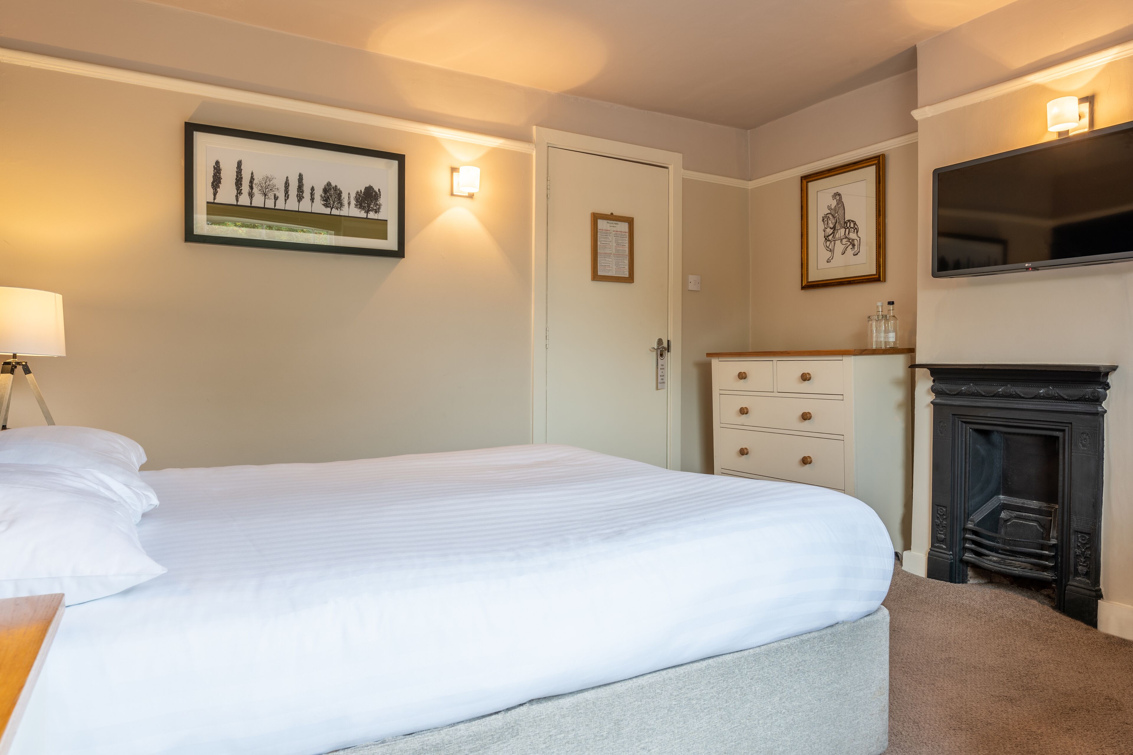 Une chambre à coucher bien rangée, avec un lit, une table de chevet, une lampe, un luminaire fixé au mur, un tableau encadré, une porte avec un avis affiché, une commode et une cheminée.