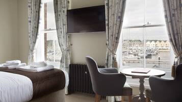A hotel room with a bed, a TV mounted on the wall, a table with chairs, and a view of a marina through large windows.