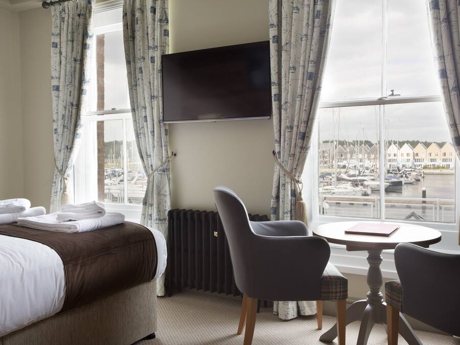 A hotel room with a bed, a TV mounted on the wall, a table with chairs, and a view of a marina through large windows.