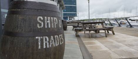 Un barril de madera con la inscripción "SHIP TRADE", una mesa de picnic y un puerto deportivo con barcos al fondo.