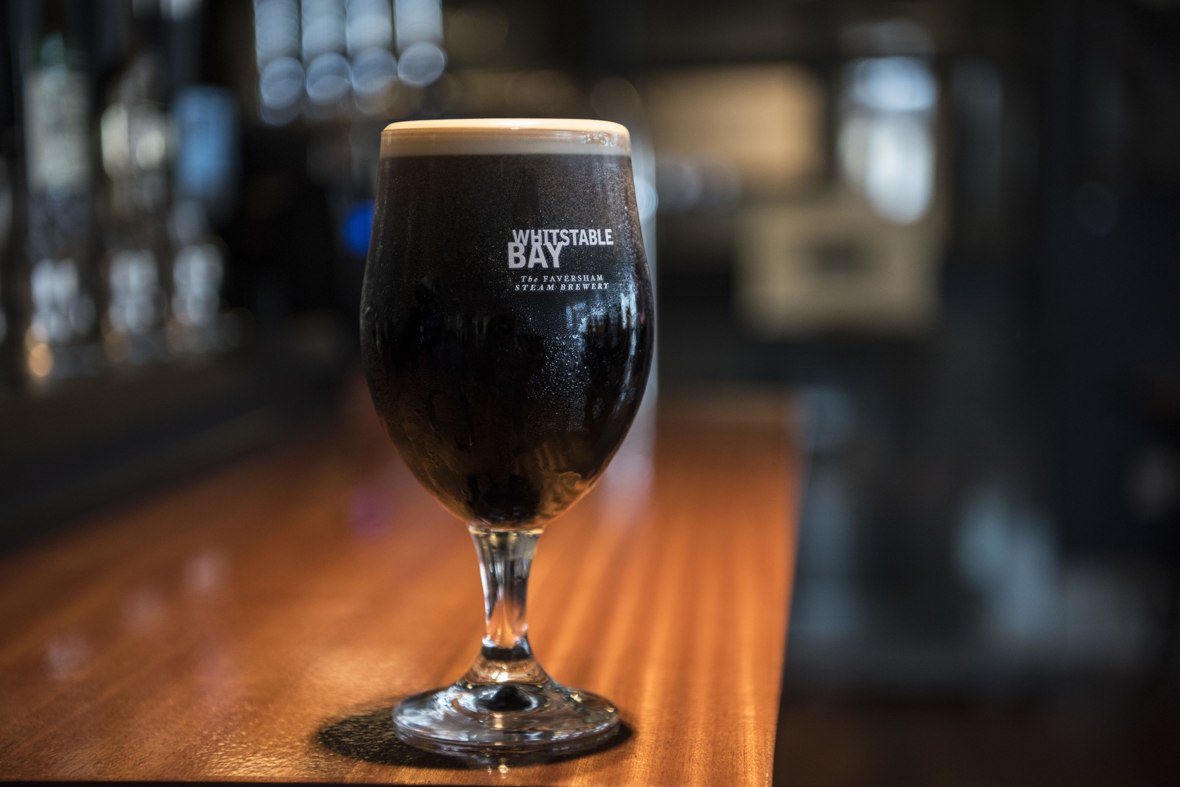 Una jarra de cerveza oscura con el texto "WHITSTABLE BAY". Está sobre una superficie de madera.