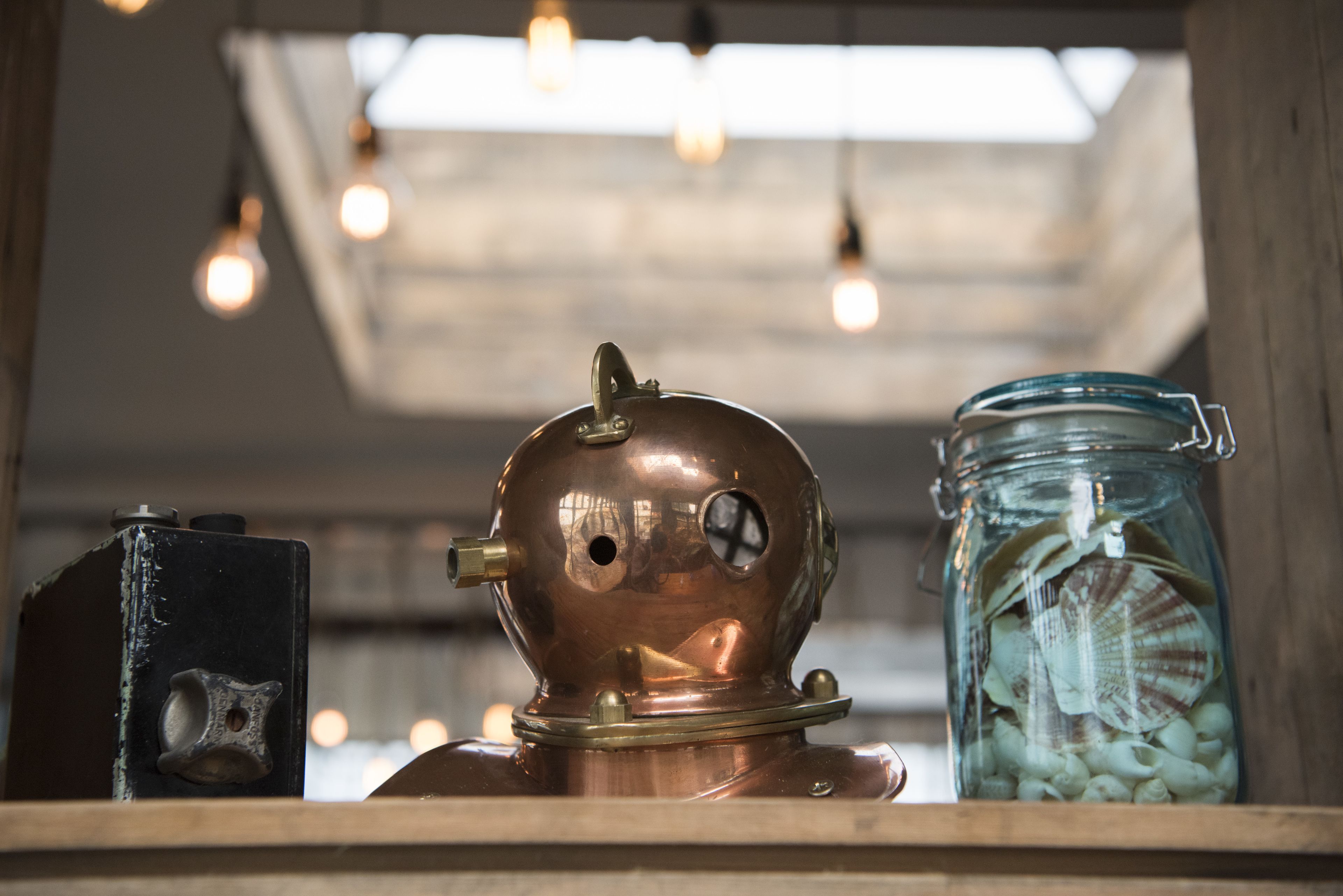 A vintage diving helmet, a glass jar with seashells, and a black object on a wooden shelf.