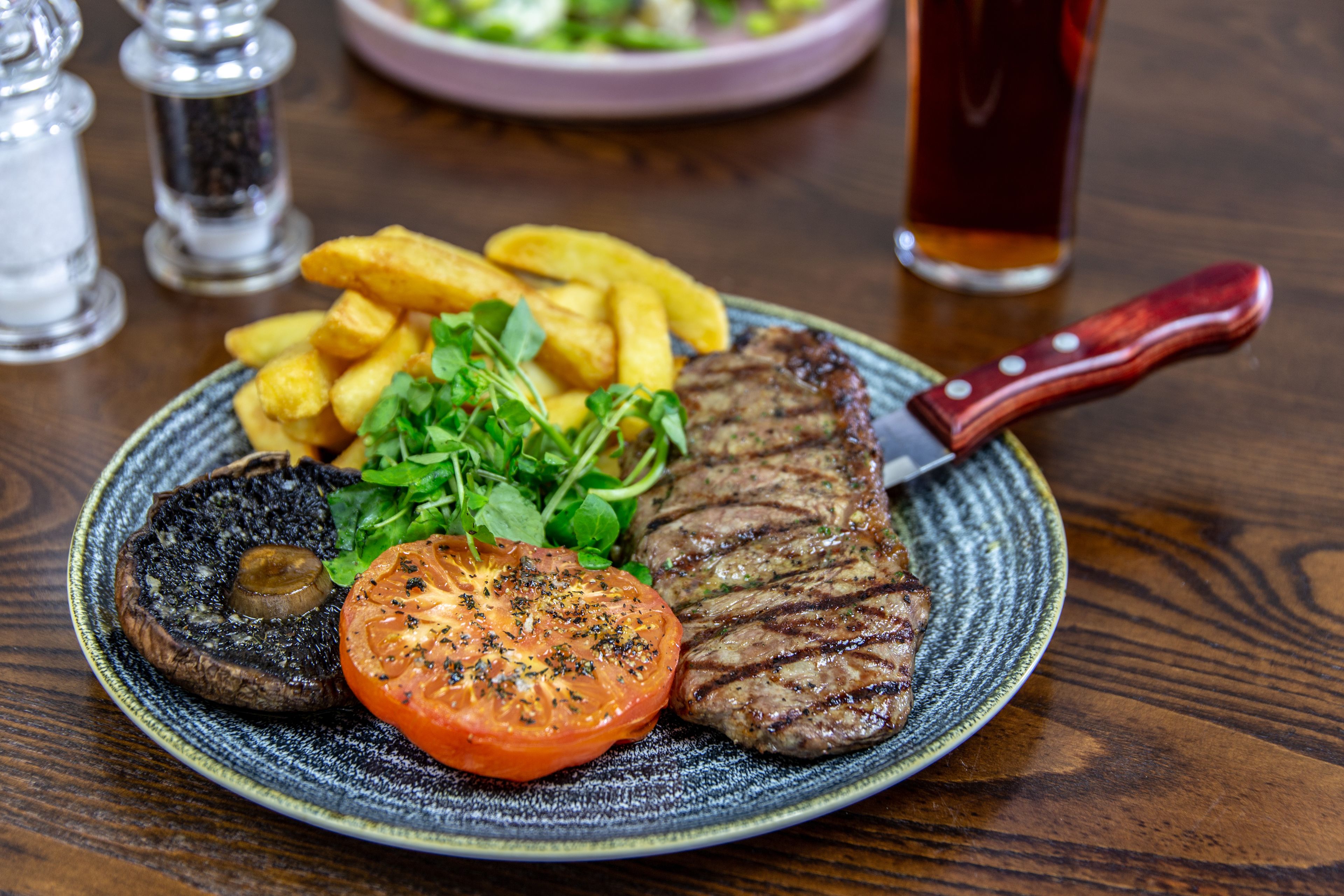 A plate with a steak, fries, a grilled mushroom, and a tomato on a ceramic plate.