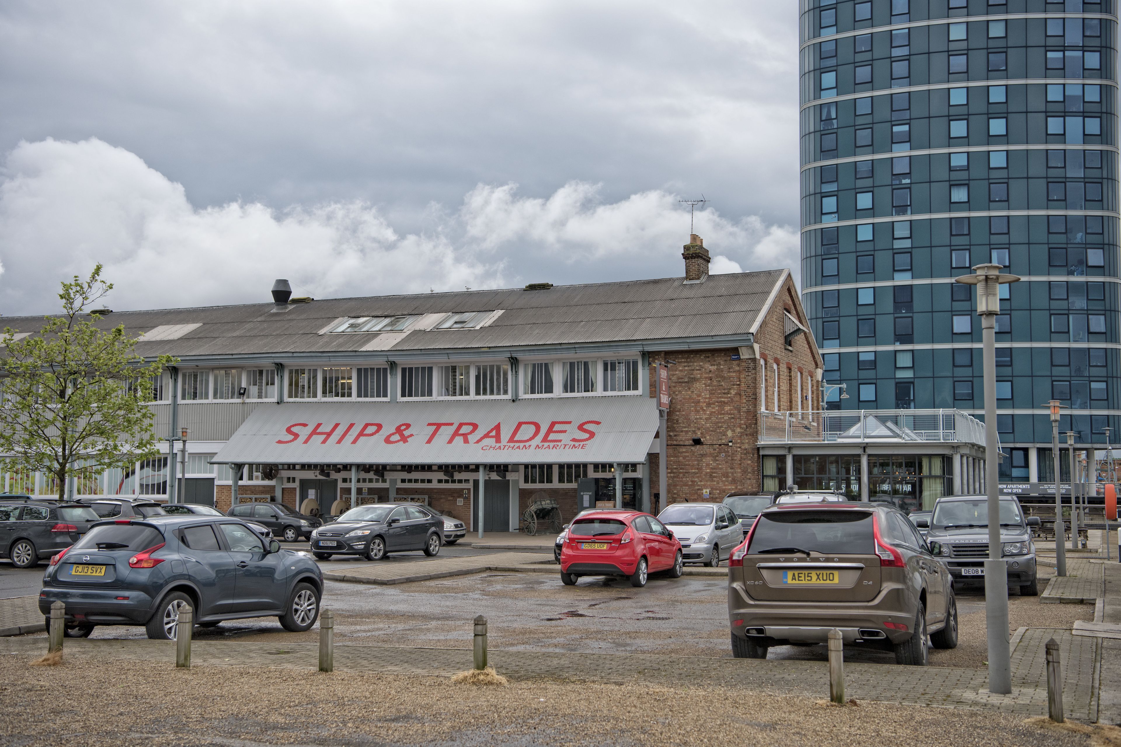 A building with a sign reading "SHIP & TRADES" and a parking lot with several cars.