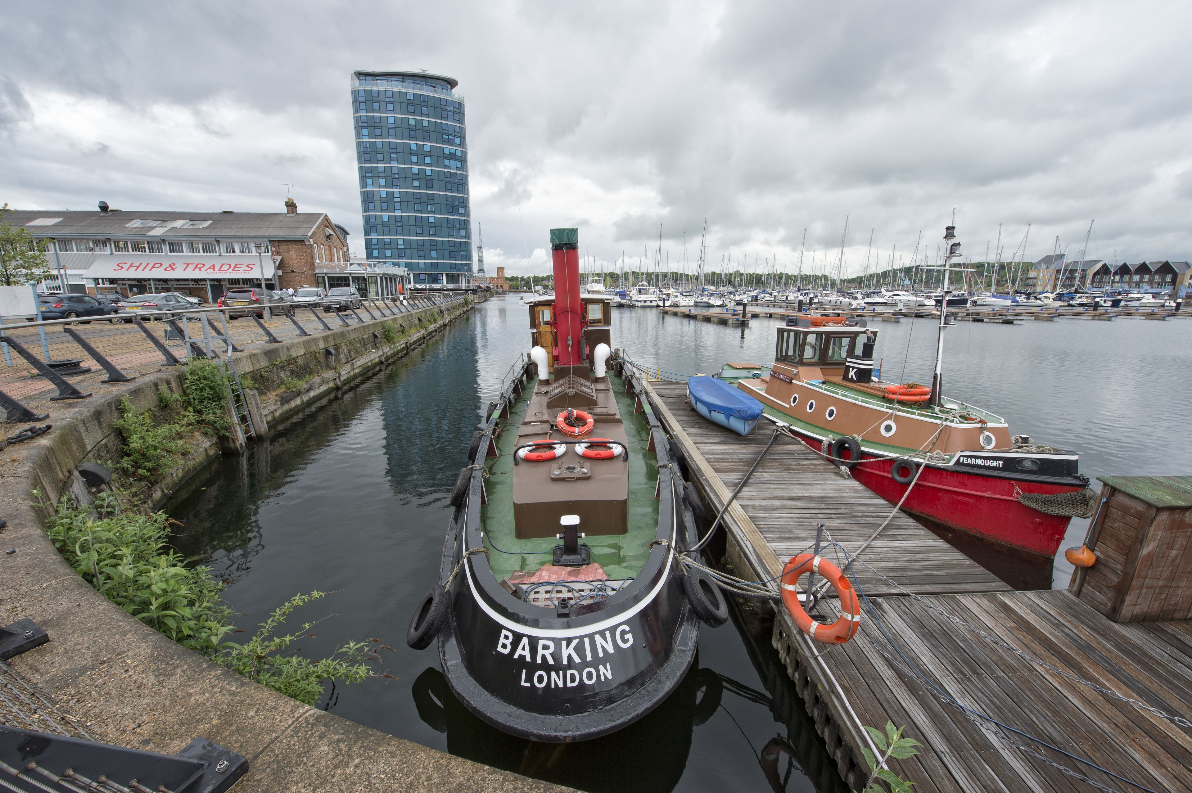 A boat docked at a marina with a building labeled "Ship & Trades" in the background.