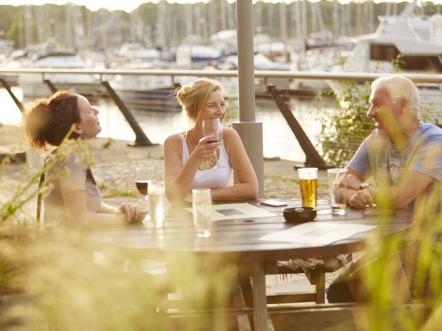 Three people sitting at an outdoor table with drinks, overlooking a marina with boats.