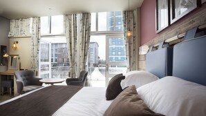 A hotel room with a large bed, a desk, a chair, and a view of the city through large windows.