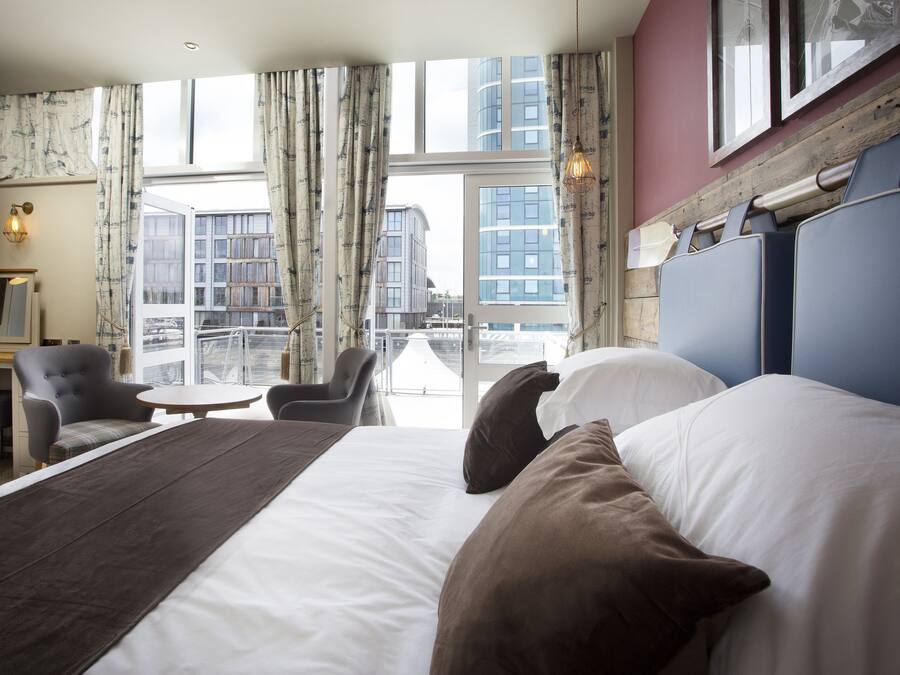 A hotel room with a large bed, a desk, a chair, and a view of the city through large windows.