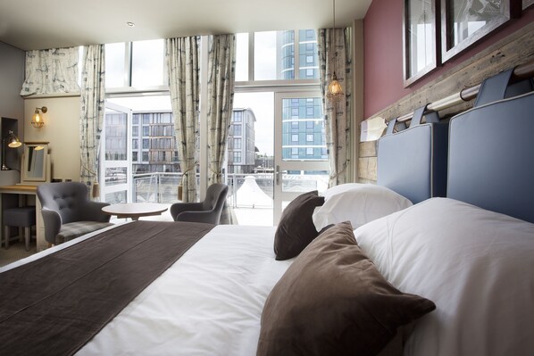 Habitación de hotel con una cama grande, un escritorio, una silla y vistas a la ciudad a través de amplios ventanales.