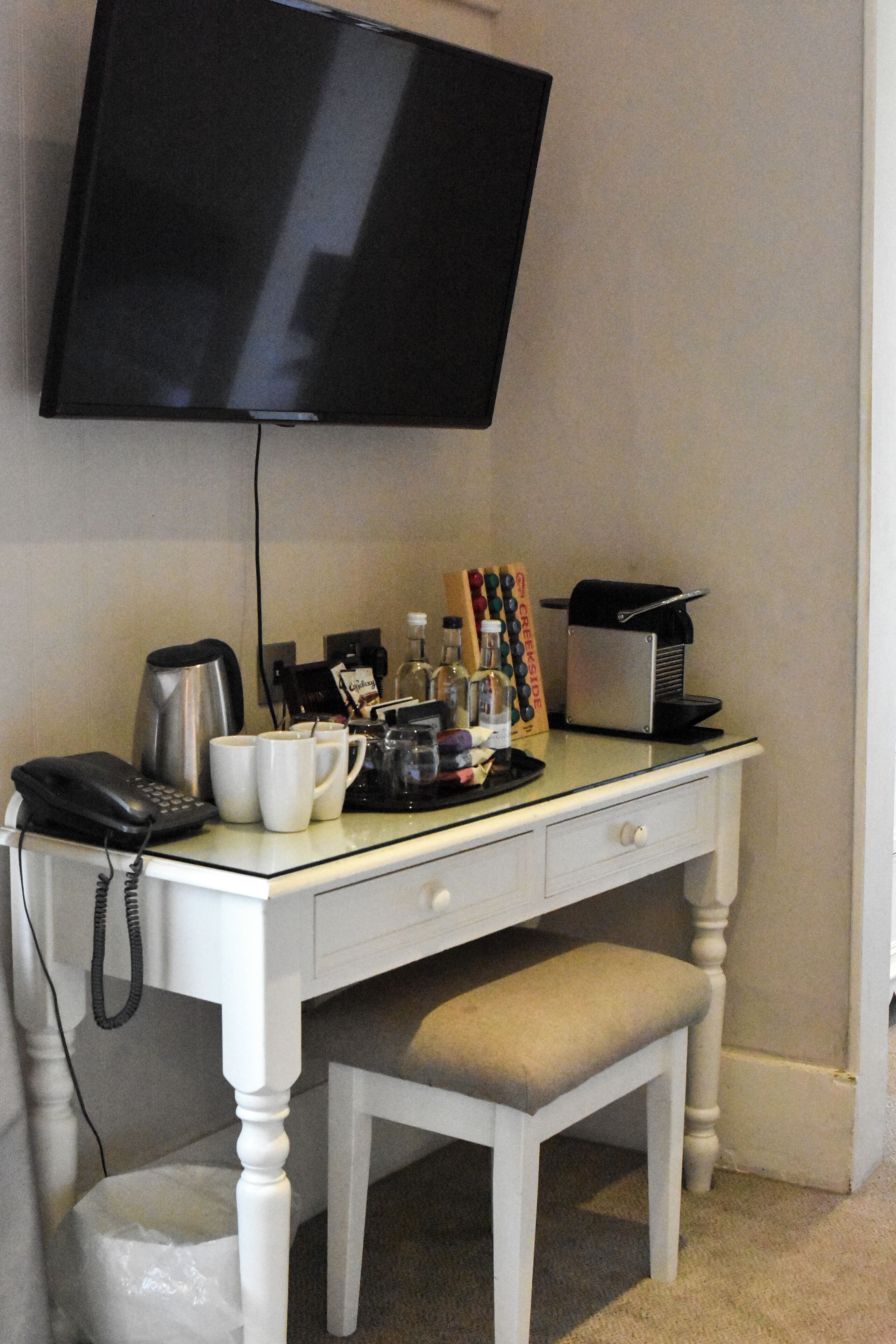 A hotel desk with a flat-screen TV, a coffee maker, mugs, a phone, and a trash bin.
