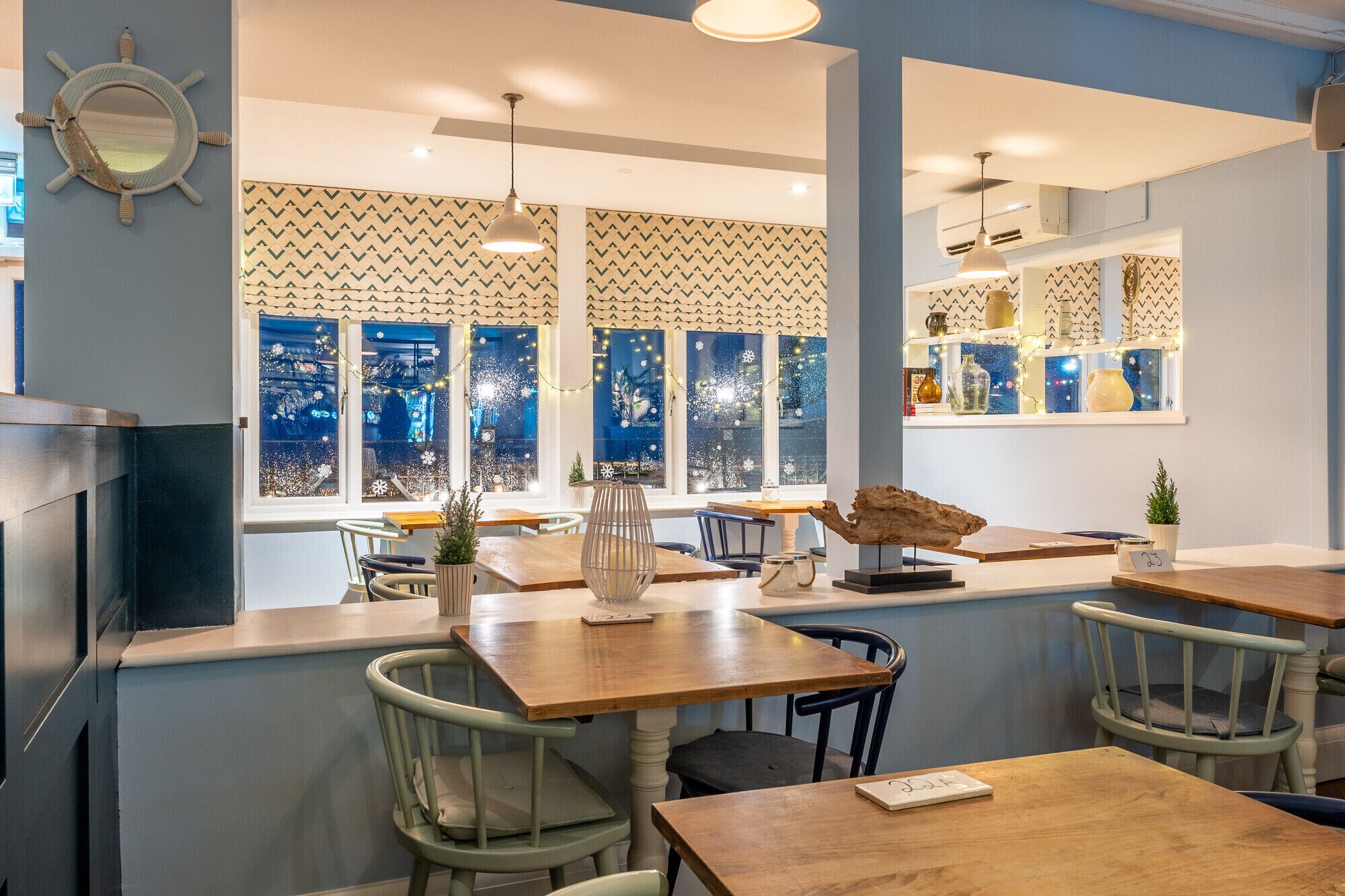 A cozy restaurant with wooden tables and chairs, a large window with a view of the city, and decorative items on the windowsill.