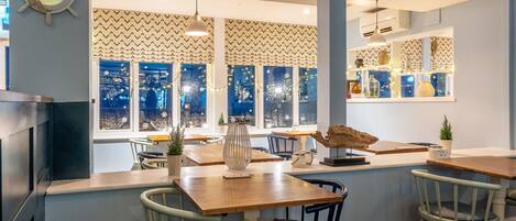 A cozy restaurant with wooden tables and chairs, a large window with a view of the city, and decorative items on the windowsill.