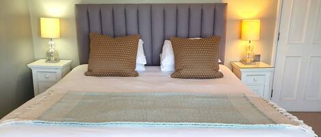 Una cama bien hecha con cabecera gris, dos lámparas de noche y una manta decorativa.