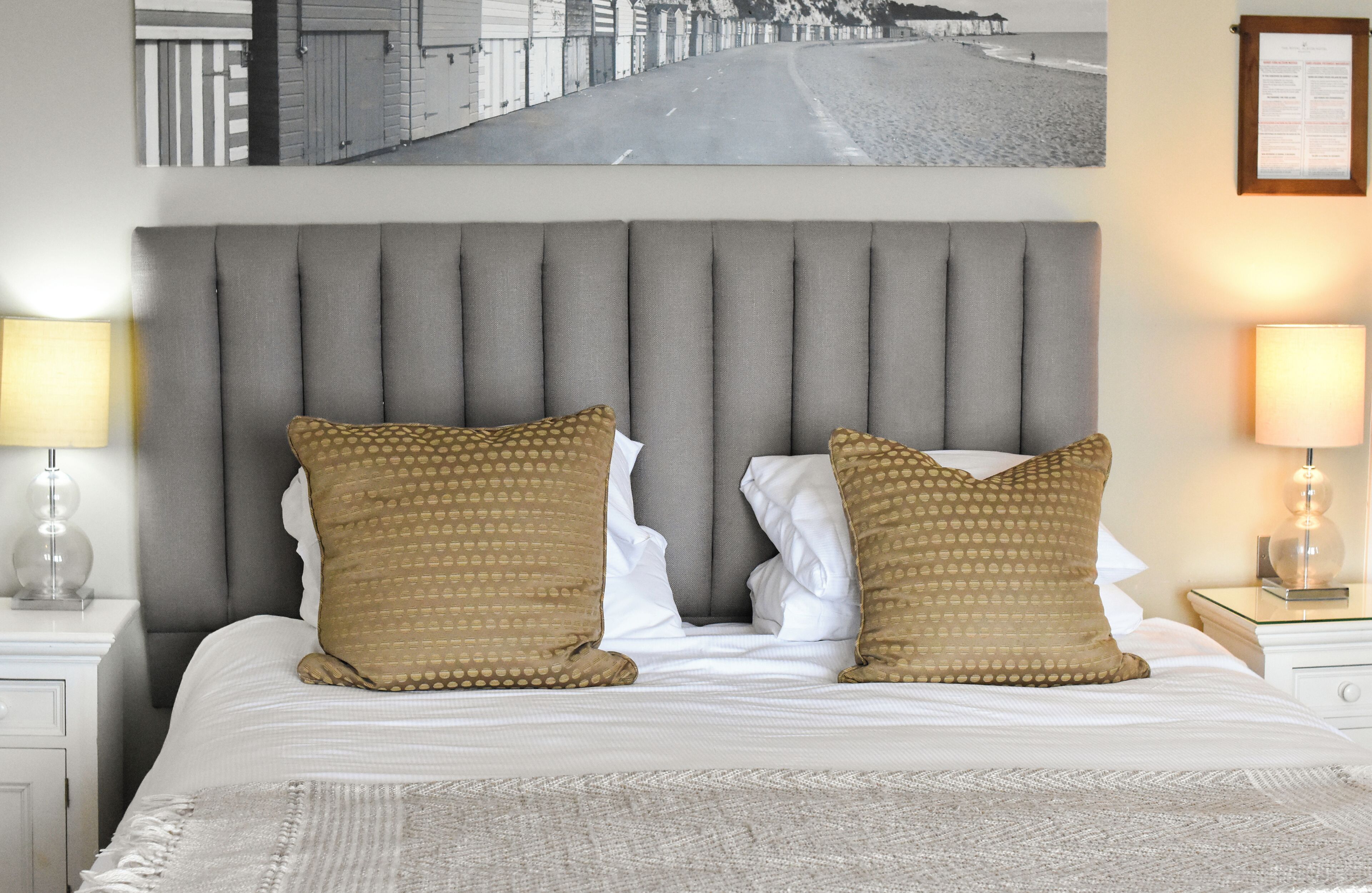 A neatly made bed with a grey upholstered headboard, two patterned pillows, and a framed picture above.