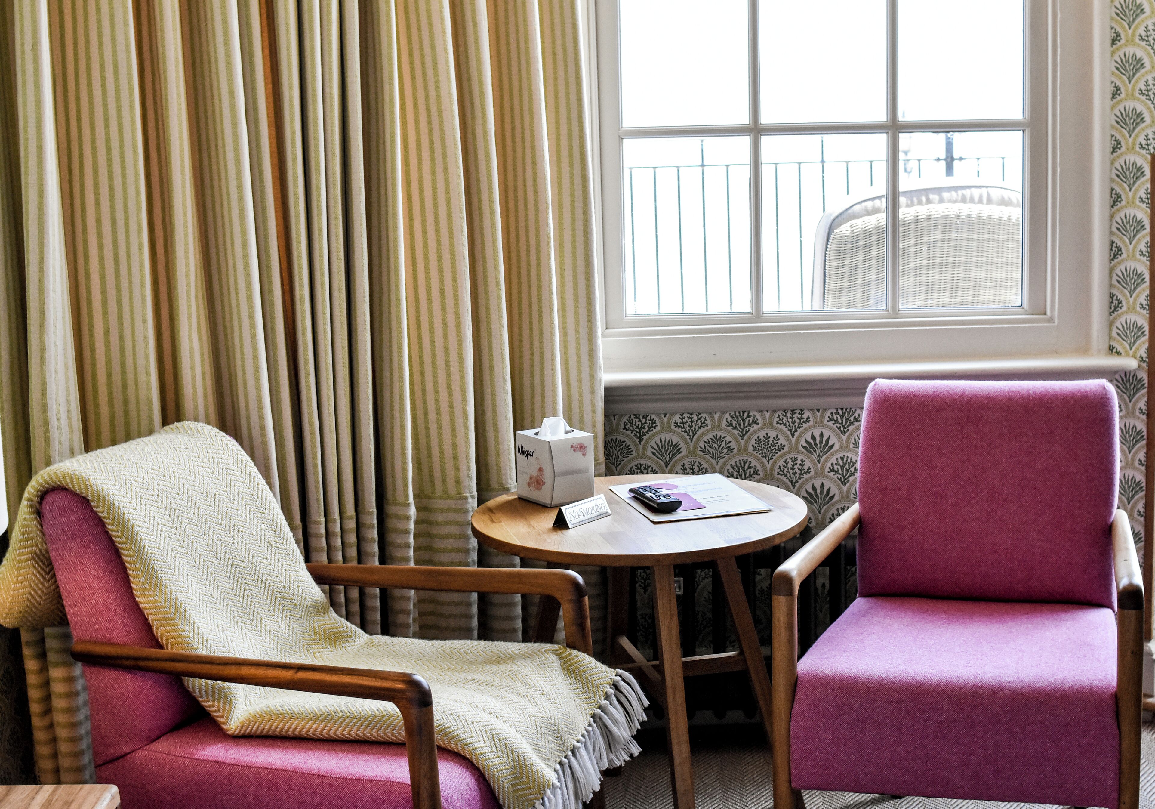 A room with a wooden chair, a pink chair, a small round table, and a window with a view of a balcony.