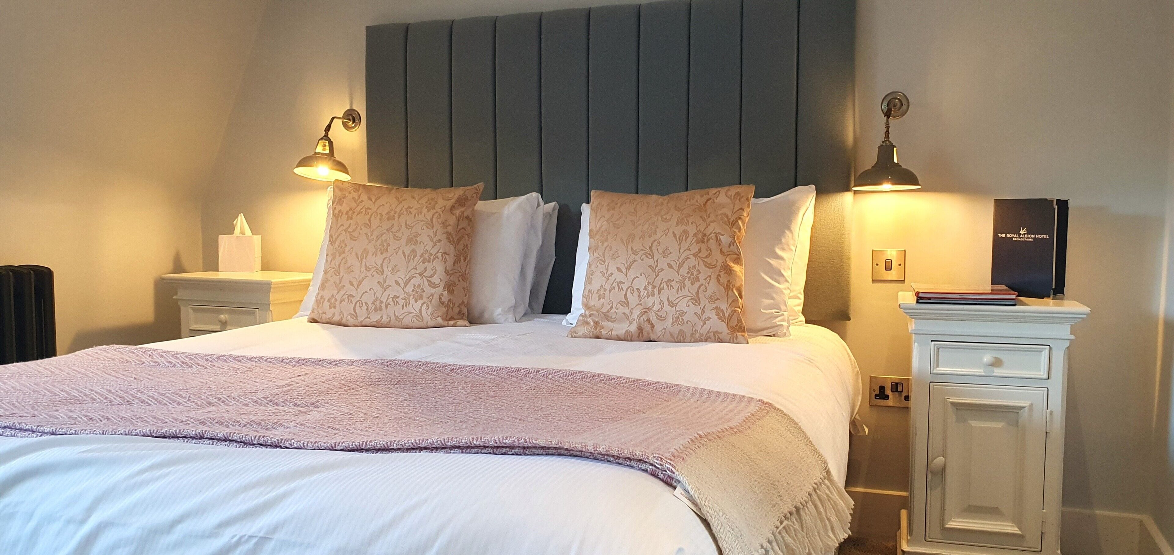 A neatly made bed with a patterned throw, two bedside lamps, and a nightstand with a book and a tissue box.