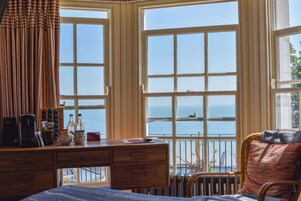 A room with a wooden desk, a bed, and large windows overlooking the sea.
