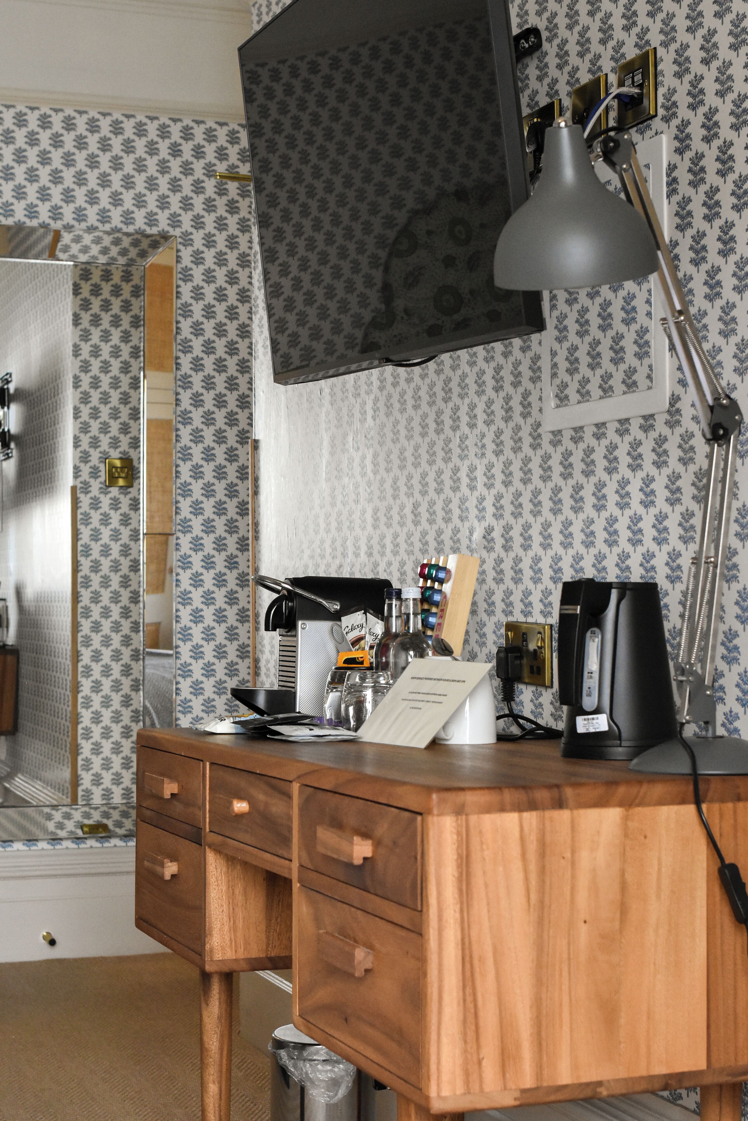 A wooden desk with a lamp, a coffee maker, and a small kitchen area with a microwave.