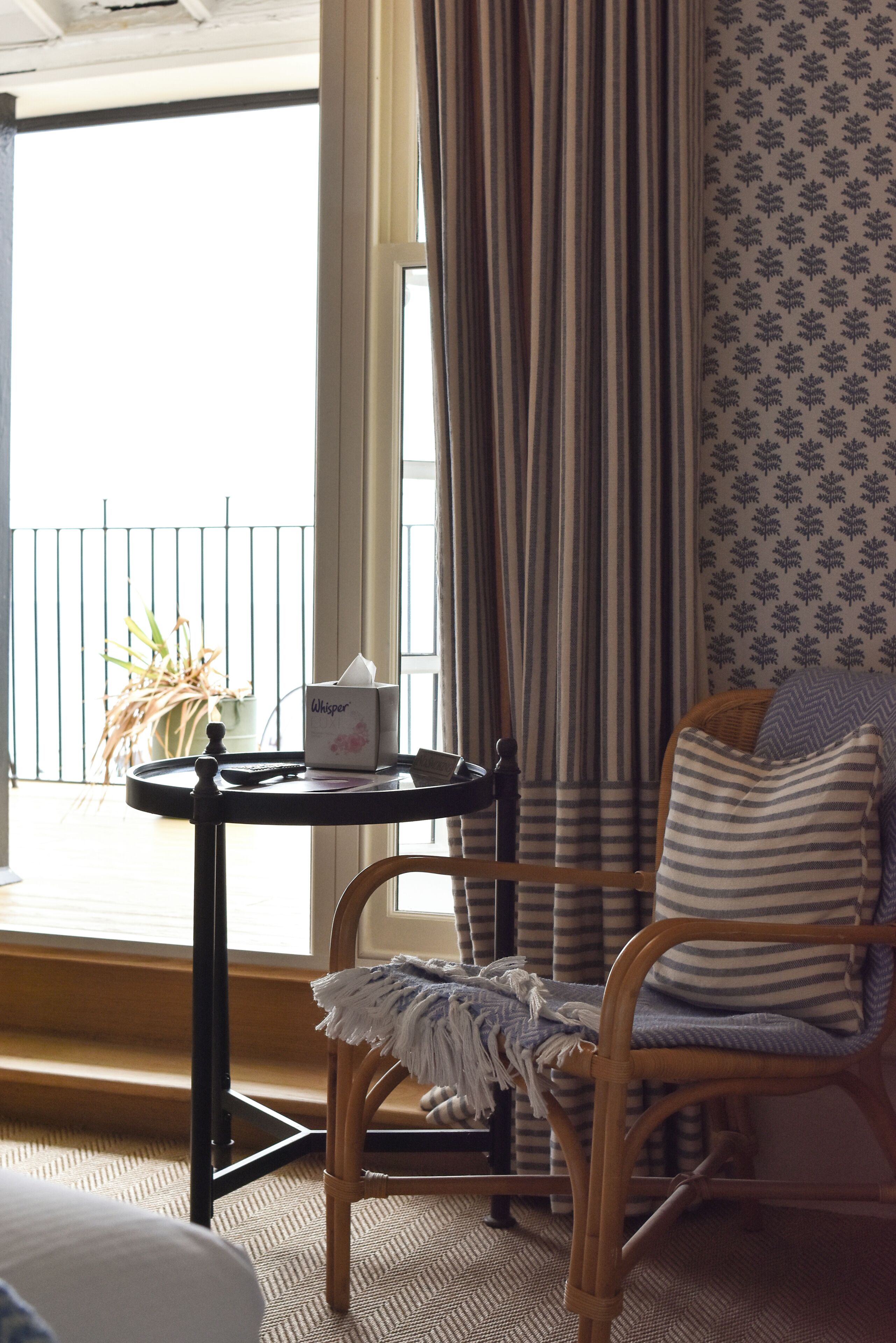 A room with a wicker chair, a small table with a tissue box, striped curtains, and a patterned wall.
