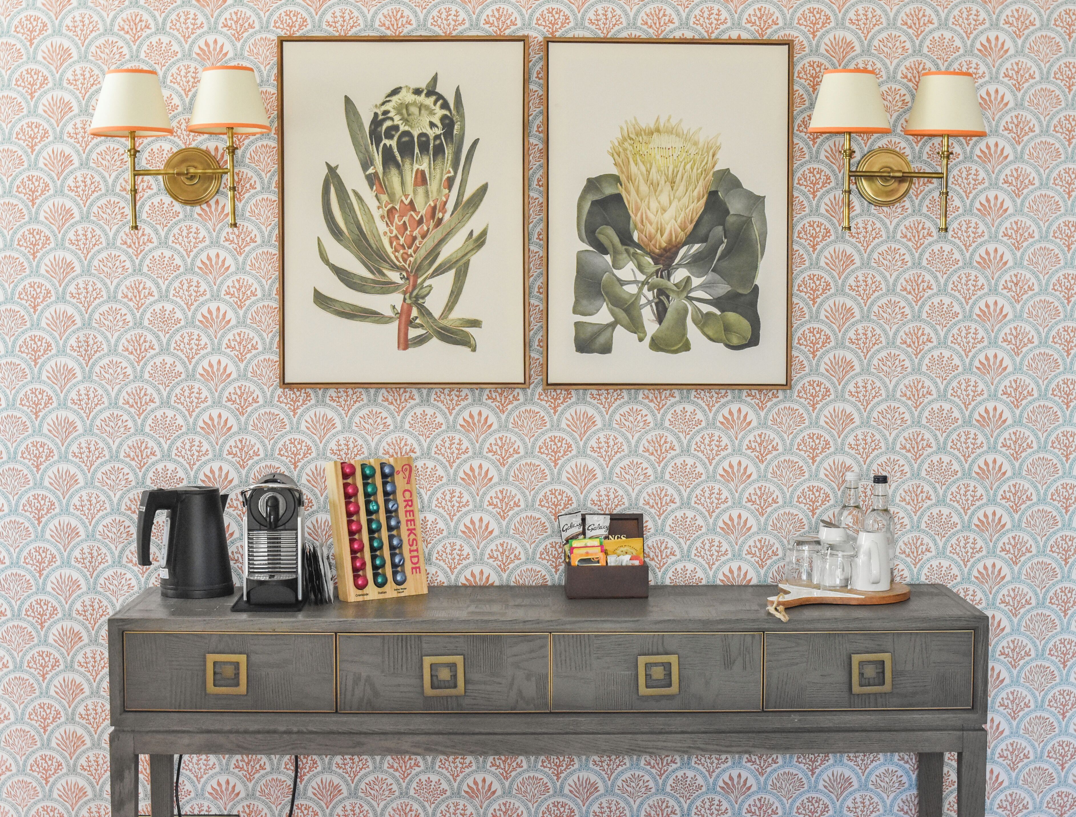 Un mur orné de deux reproductions botaniques encadrées, une machine à café, un ensemble comprenant un pot à crème et un sucrier, et un plateau avec des verres et une théière, le tout posé sur une console en bois.
