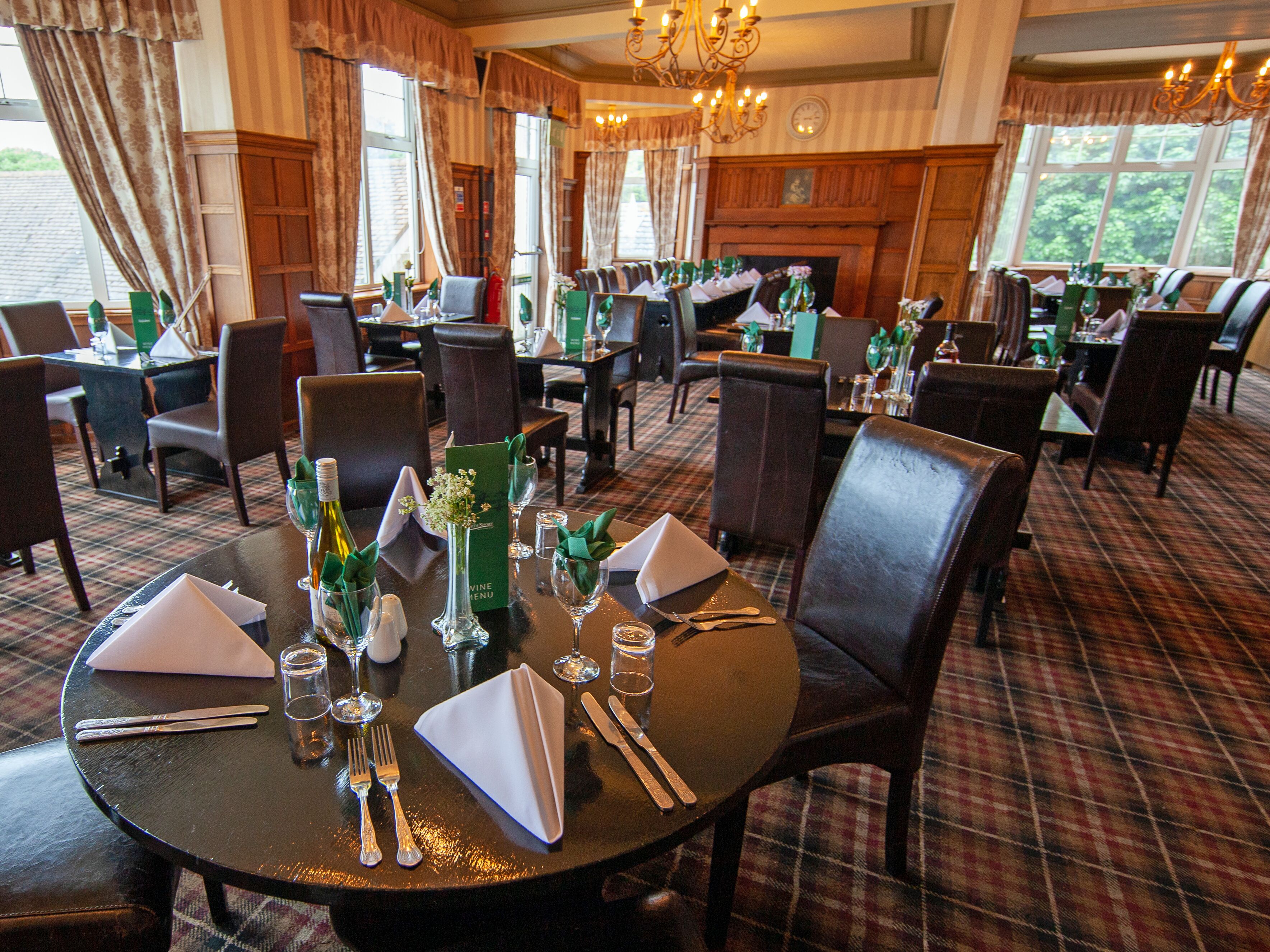 A dining area with round tables set for four, each with a menu, wine glass, and cutlery. The room has a plaid carpet, wooden paneling, and large windows with curtains.