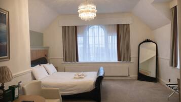 Habitación de hotel con cama, sillón, mesa, un ventanal con cortinas, candelabro y espejo.