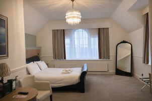 Habitación de hotel con cama, sillón, mesa, un ventanal con cortinas, candelabro y espejo.