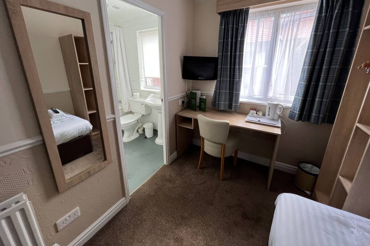 Ein Hotelzimmer mit Bett, Schreibtisch mit Stuhl, Fernseher, Fenster mit Vorhängen und einem durchgehenden Türspalt sichtbaren Badezimmer.