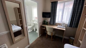 A hotel room with a bed, a desk with a chair, a television, a window with curtains, and a bathroom visible through an open door.
