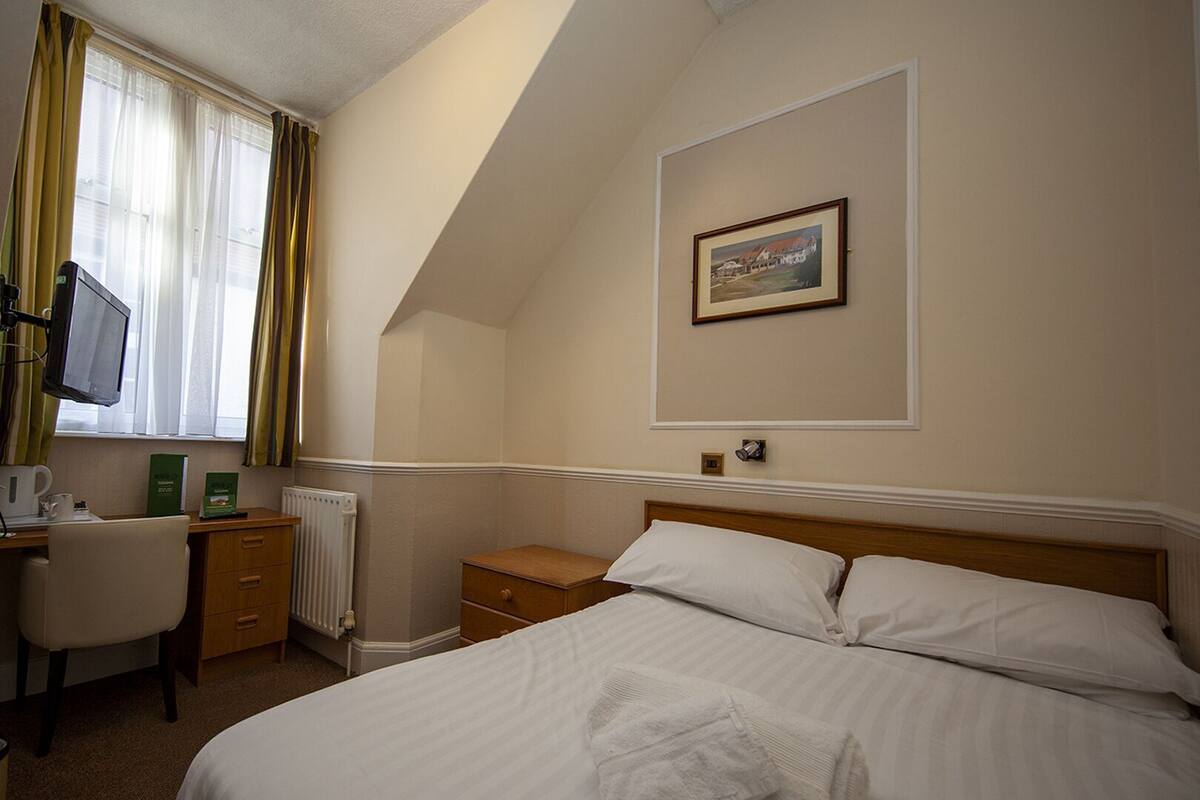 Ein Hotelzimmer mit einem Bett, einem Schreibtisch mit Computer, einem Sessel, einer Heizung und einem gerahmten Bild an der Wand.