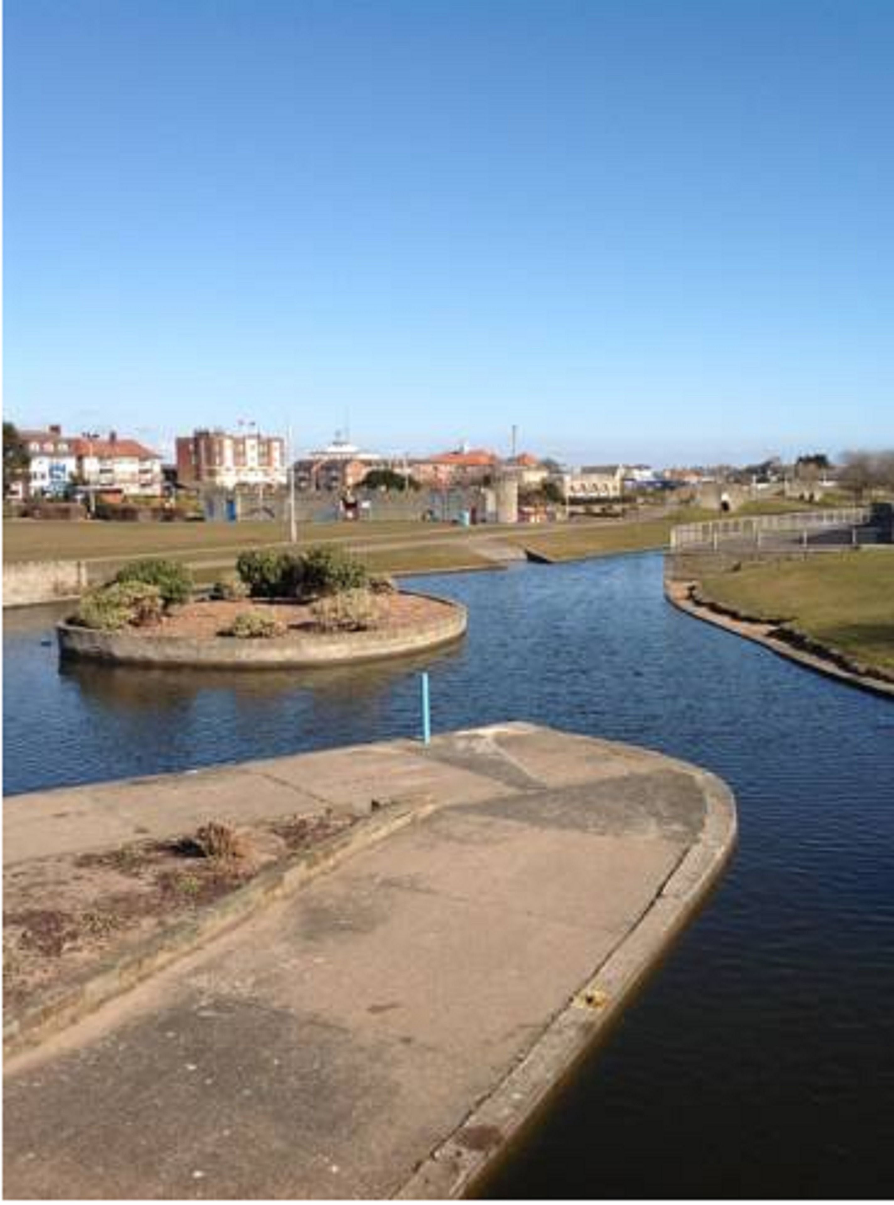 Un canal con una isla circular, edificios al fondo y un cielo despejado.