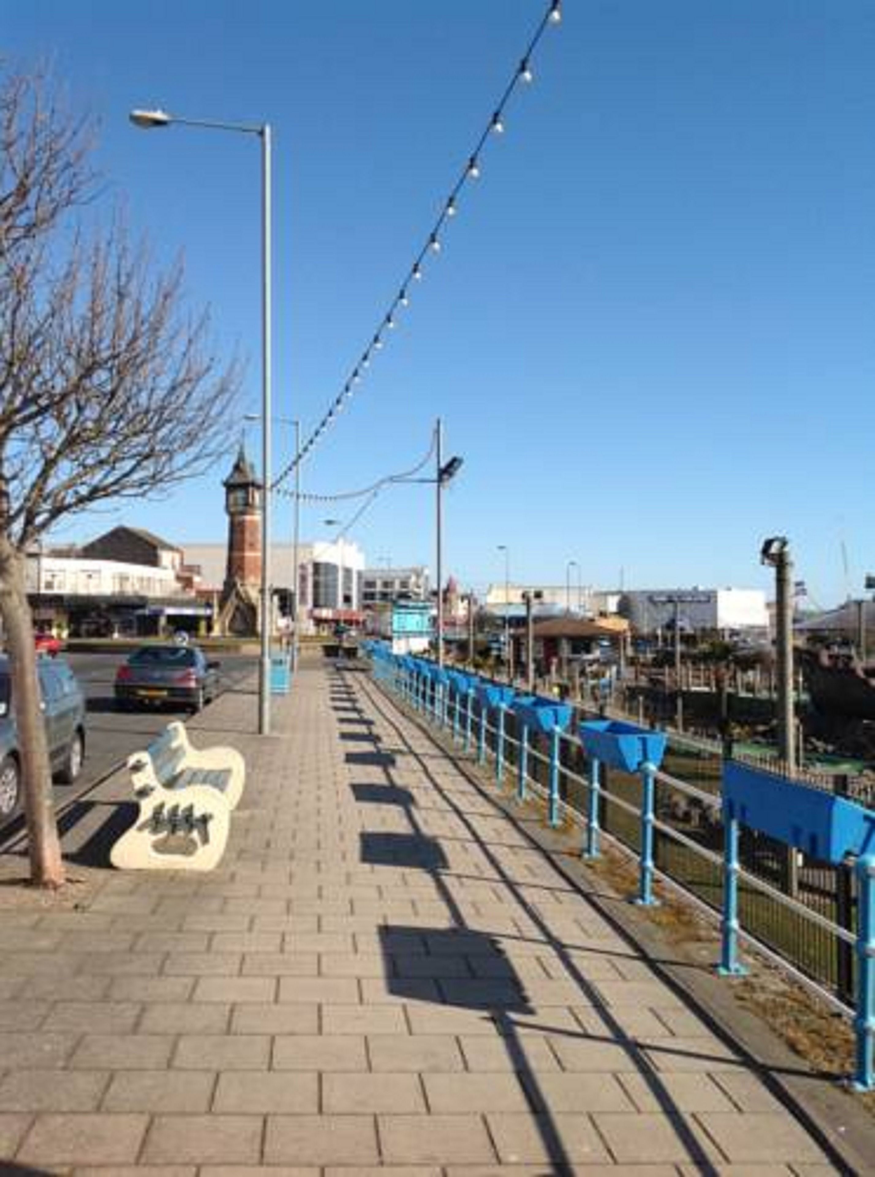 A paved walkway with a blue railing, a street lamp, and a building with a tower in the background.