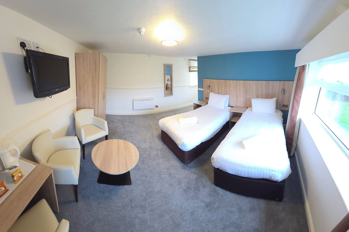 Ein Hotelzimmer mit zwei Einzelbetten, einem Schreibtisch mit Stuhl, einem kleinen runden Tisch, einem an der Wand montierten Fernseher und einem Fenster mit Vorhängen.