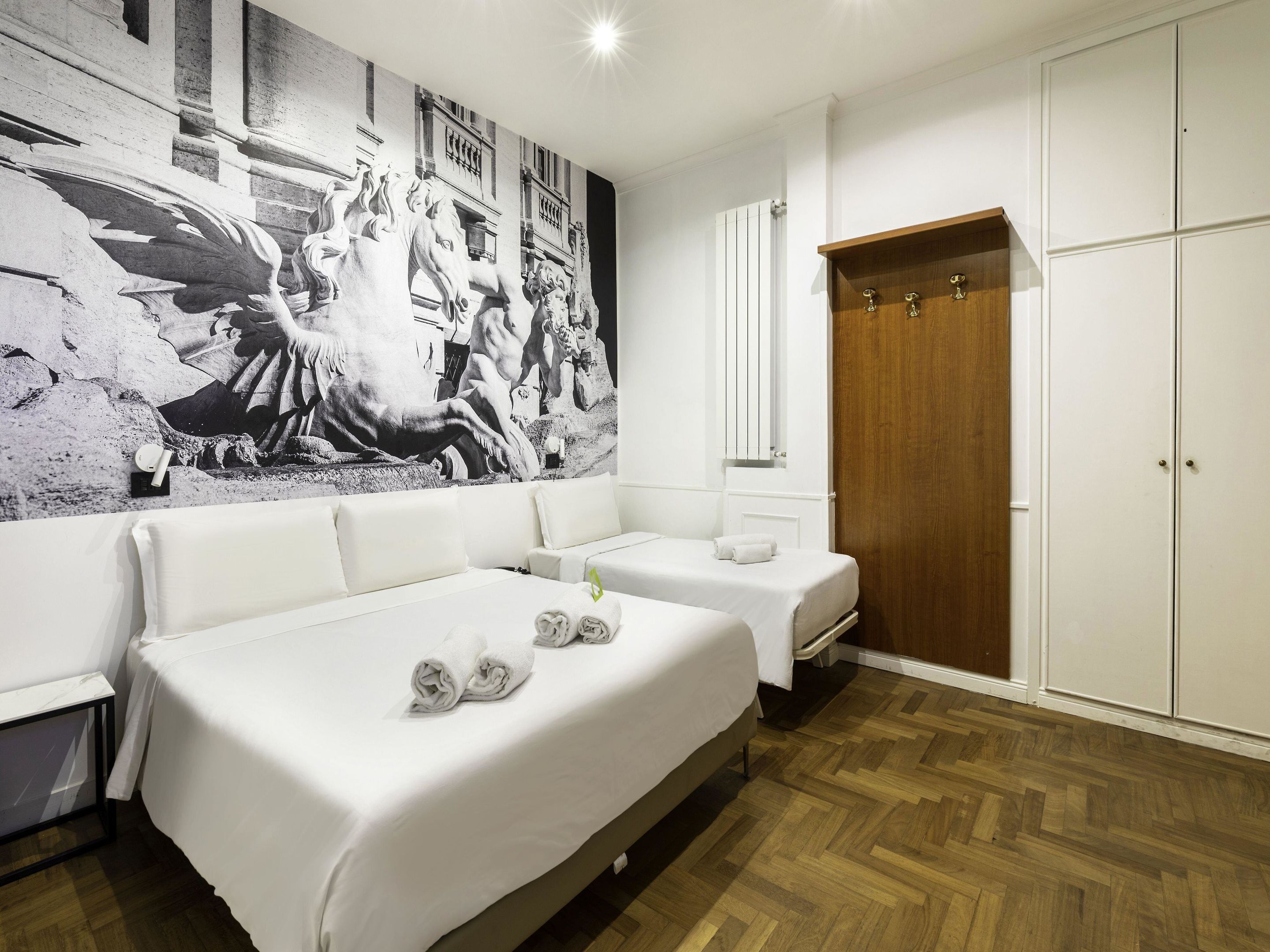 Una camera d'albergo con un grande murale in bianco e nero, un armadio in legno, due letti e un comodino.