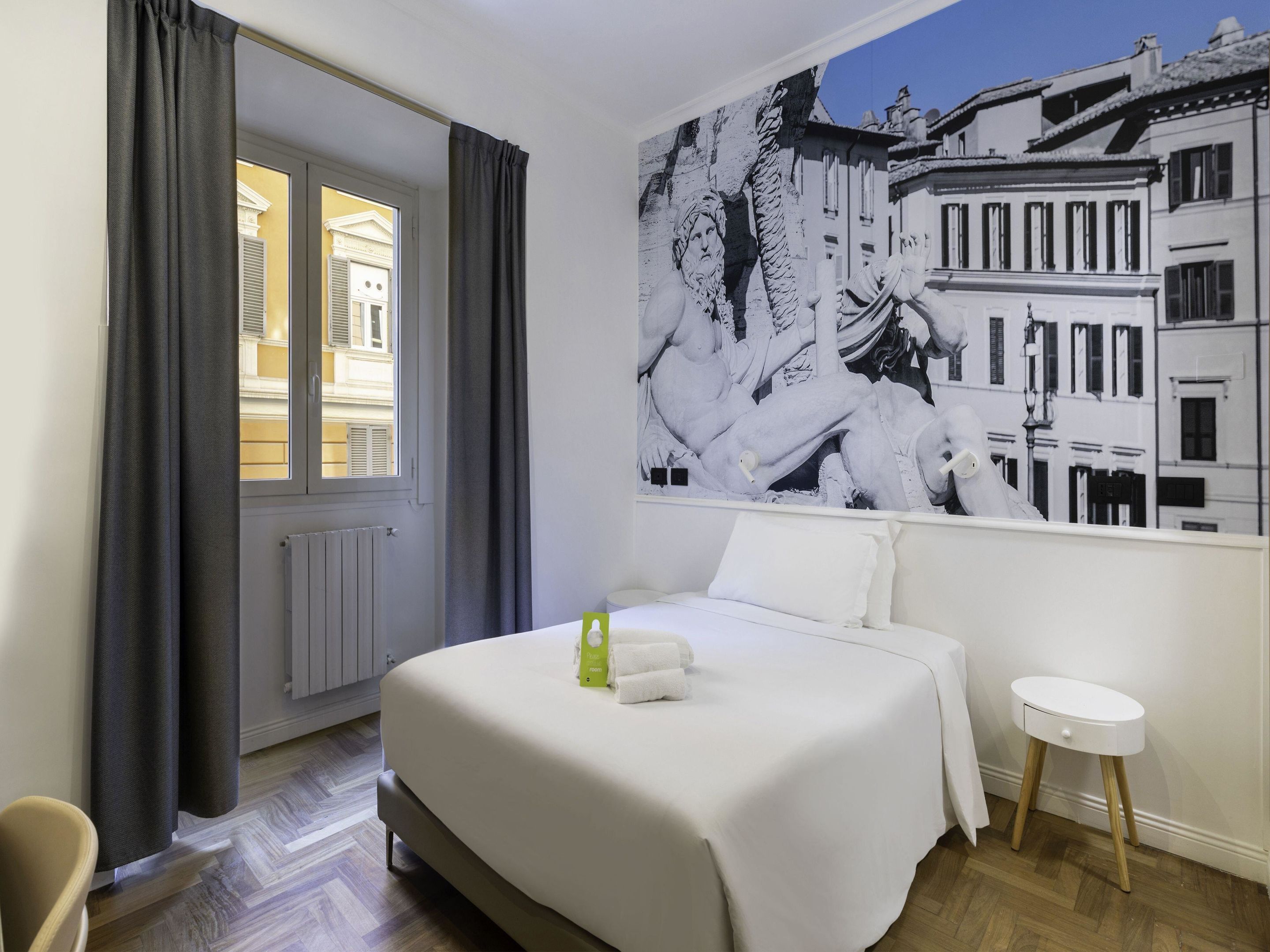 A hotel room with a bed, a window with curtains, a radiator, a small stool, and a wall with a black and white cityscape mural.
