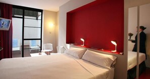 Una habitación de hotel con una cama grande, una pared de acento roja, dos lámparas de noche, una pequeña mesa con una silla y vistas a través de un ventanal amplio.
