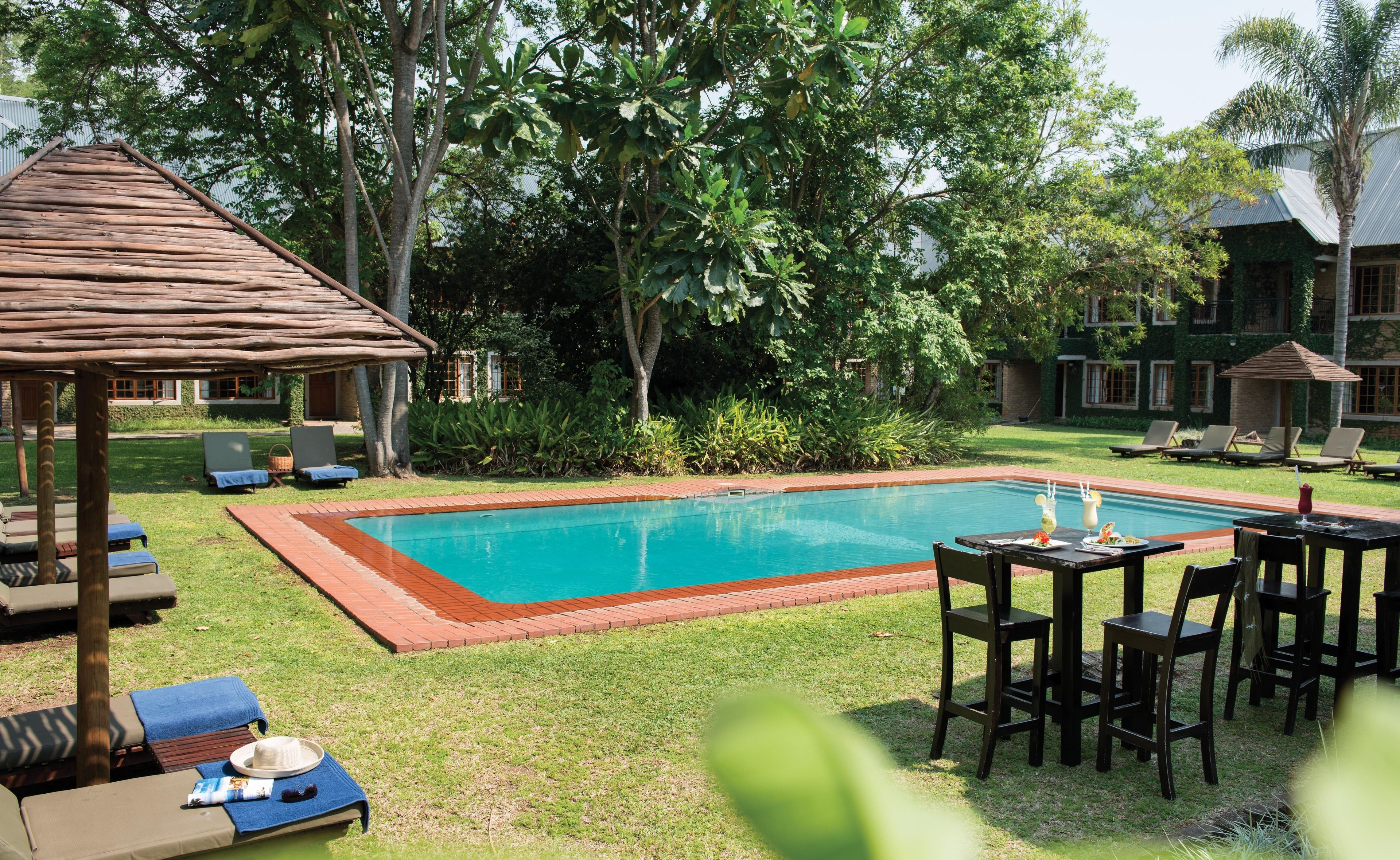 Un espace piscine avec des chaises longues et une petite table où sont servis des boissons, le tout entouré de verdure et avec un bâtiment en arrière-plan.