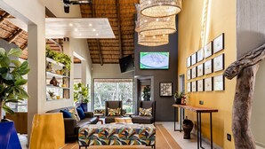 A spacious living room with a large sofa, two armchairs, a coffee table, and a TV mounted on the wall. There are multiple potted plants and a large window providing natural light. - Hippo Hollow Country Estate (Mbombela)