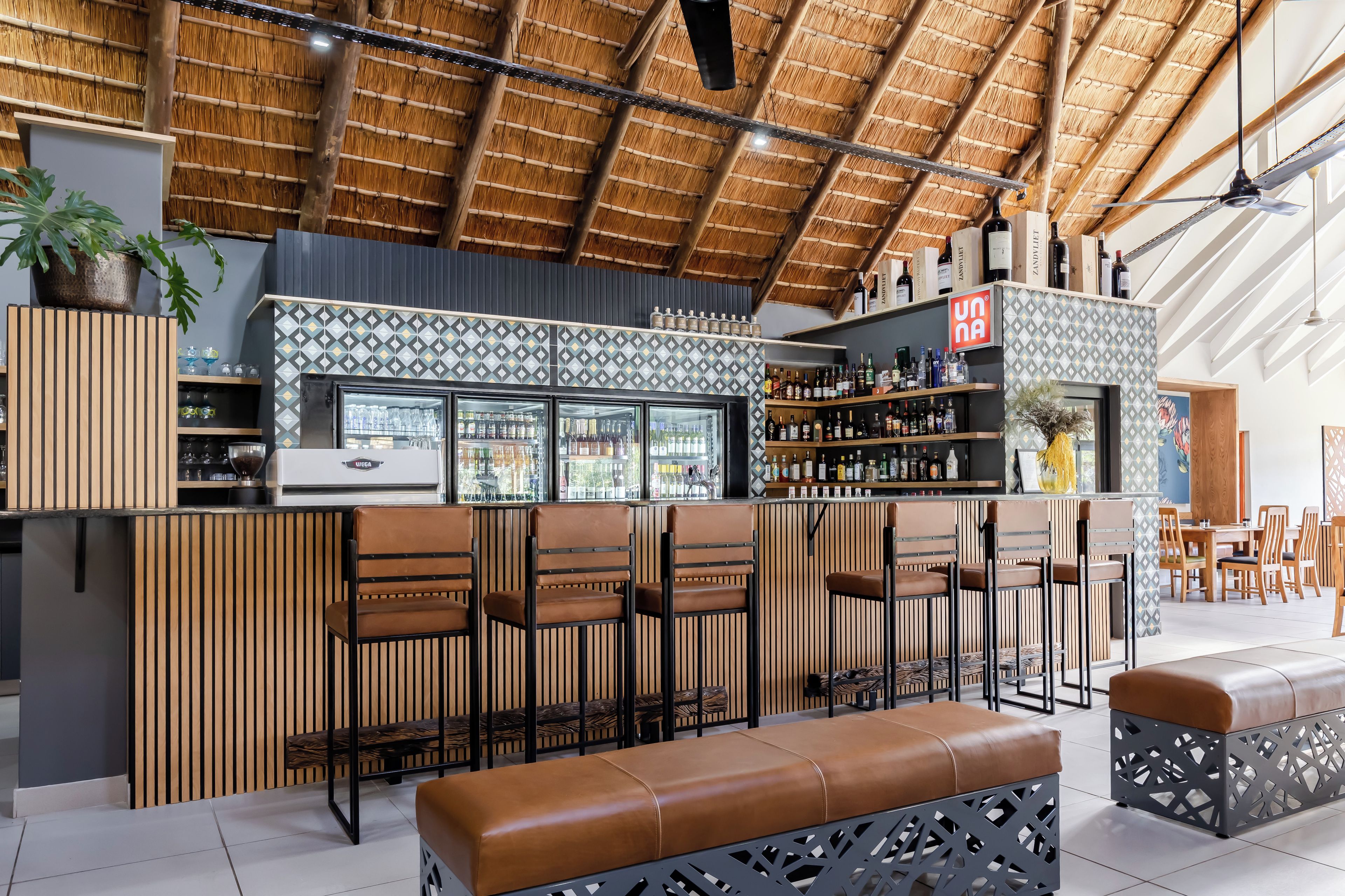 A bar area with a thatched roof, wooden slatted walls, and a variety of bottles and glasses on shelves.