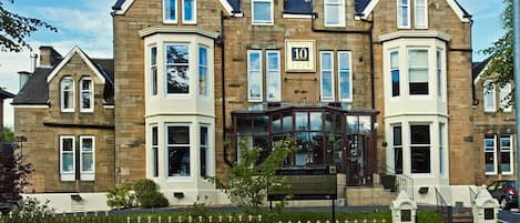 A large, two-story stone building with a prominent bay window and a sign indicating it is a hotel.