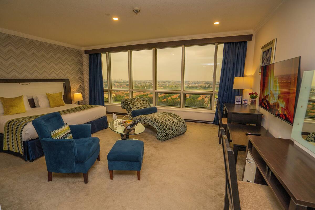 A hotel room with a large bed, a blue armchair, a matching ottoman, a small round table, and a TV.