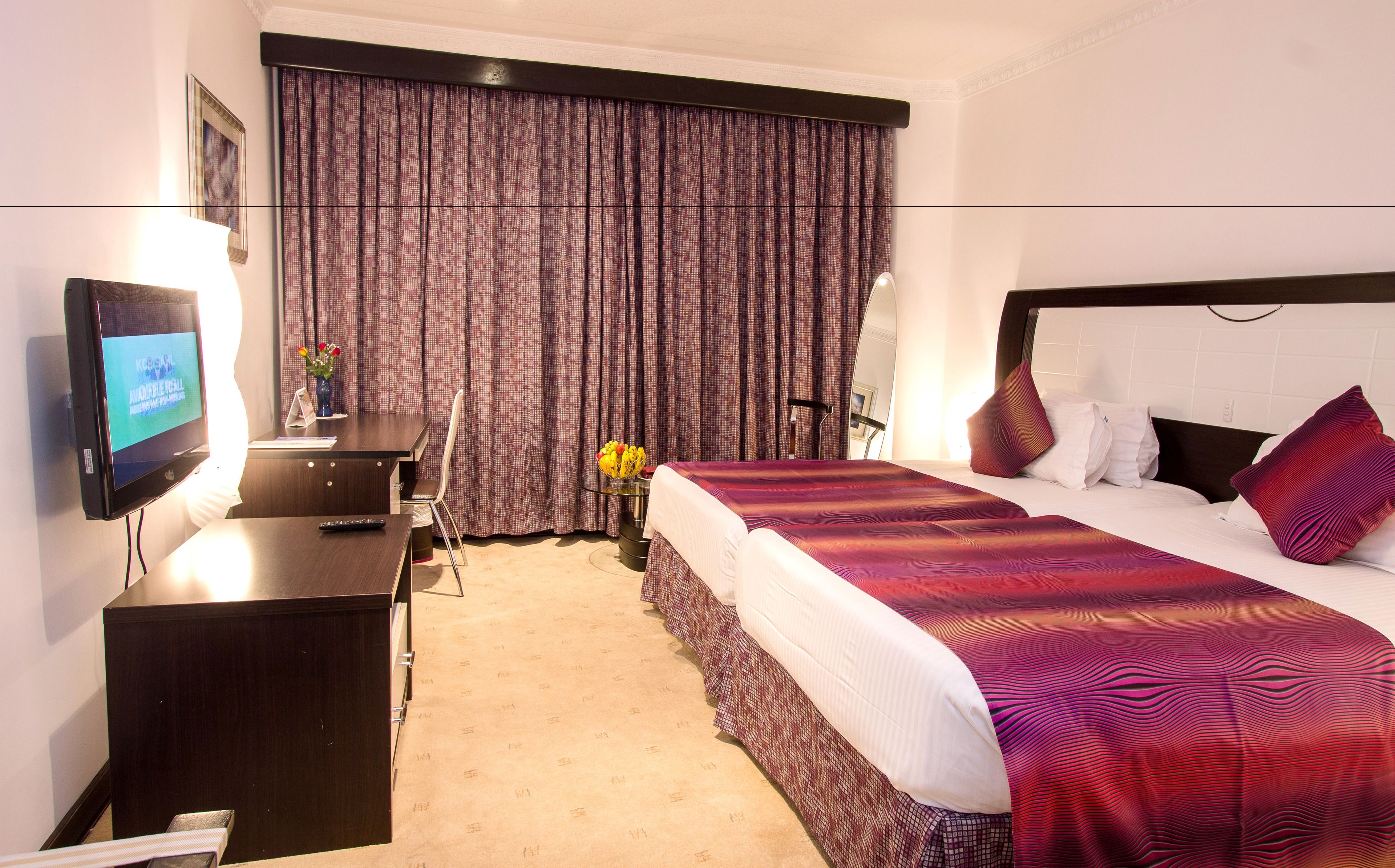 Habitación de hotel con cama, escritorio, computadora, televisor y ventana con cortinas.