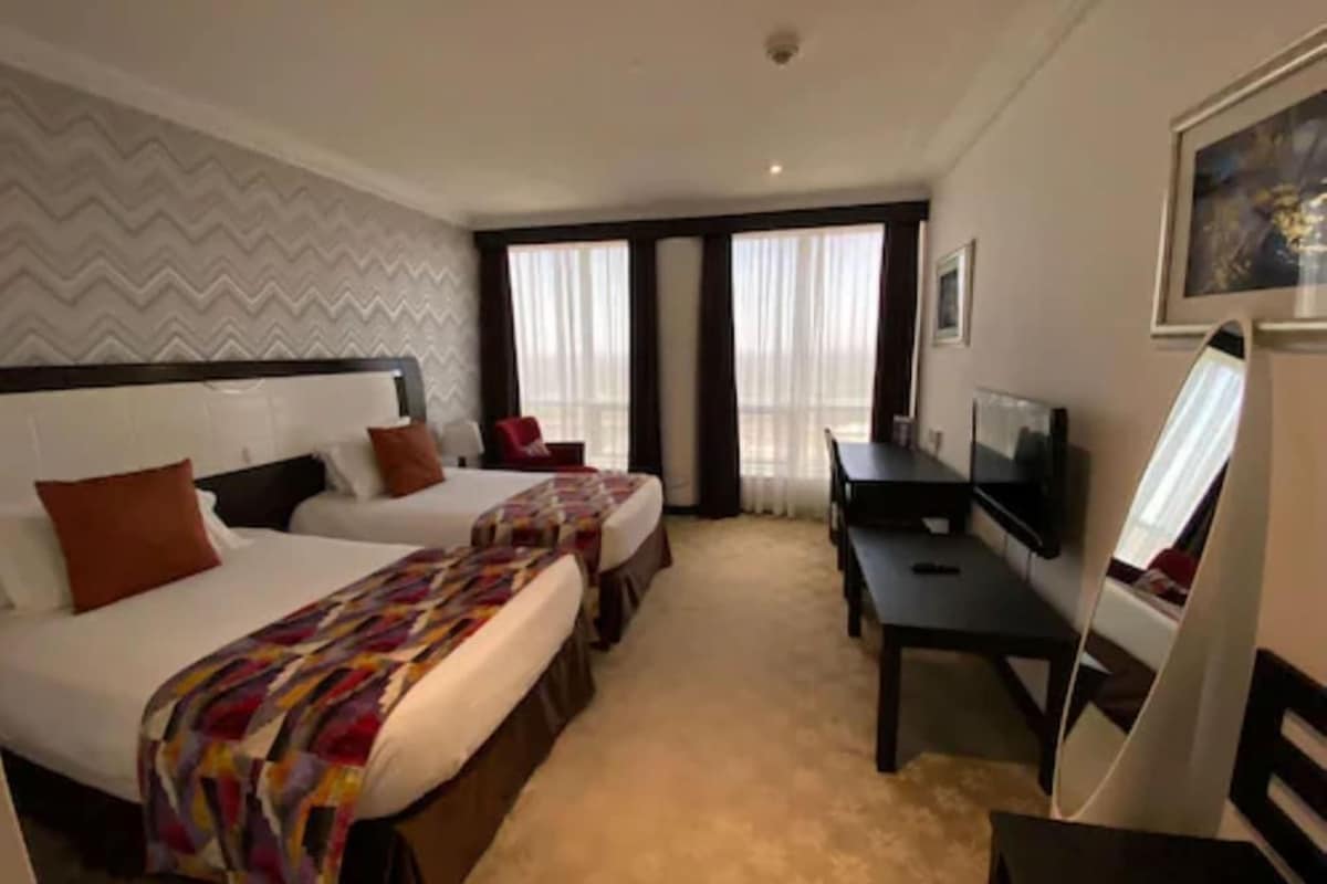 A hotel room with two beds, a desk, a TV, and a large window.
